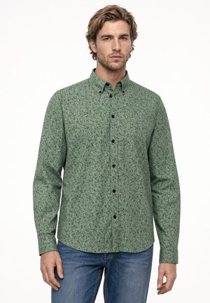 Hombre con una camisa verde de manga larga con patrón de cuadros negros, combinada con vaqueros azules, de pie frente a un fondo liso.