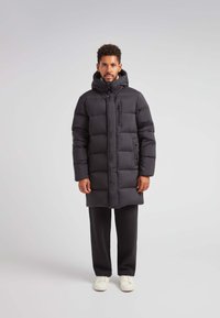 Czarna puchowa kurtka z kapturem, z zamykanym na zamek kieszonką na piersi, z zatrzaskami oraz pikowanym wzorem dla lepszej izolacji. Noszona na czarnych spodniach.