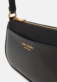 Sac bandoulière noir en matériau texturé avec une fermeture éclair dorée et un logo. Le sac a une forme arrondie et élégante, avec une sangle pour le porter.