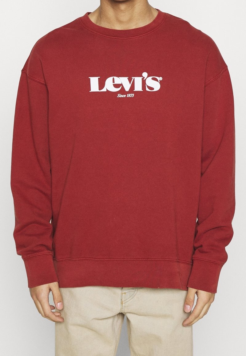 Personne portant un sweat-shirt rouge Levi's avec un logo blanc, associé à un pantalon beige clair, debout devant un fond uni.