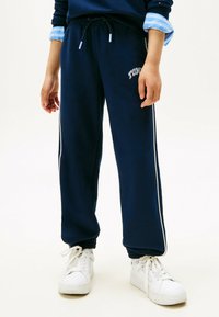 Persoon in een marineblauwe trainingsbroek met witte zijstrepen en witte sneakers, staand tegen een effen lichte achtergrond.