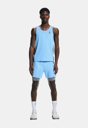 Mandlig atlet stående iført lyseblå løbetop uden ærmer og shorts med grå kompressionsshorts under, hvide sokker og løbesko.
