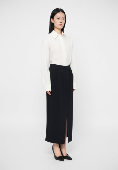 Γυναίκα όρθια φορώντας λευκό πουκάμισο μ�ε κουμπιά, μαύρη ψηλόμεση φούστα με σχισμή μπροστά, και μαύρες μυτερές γόβες με ψηλό τακούνι, στο φόντο ενός λευκού τοίχου.