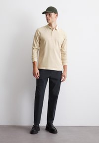 Bežna polo majica s dolgimi rokavi in ovratnikom, v kombinaciji z črnimi hlačami in črnimi čevlji na vezalke. Model nosi zeleno kapo. Minimalističen dizajn.
