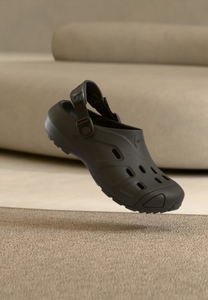 Čierne nazúvacie dreváky s ventilačnými otvormi a nastaviteľným pásikom na päte, ktoré plávajú nad textúrovaným béžovým kobercom s béžovou pohovkou v pozadí.