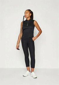 Melna bez piedurknēm polo krekls ar rāvējslēdzēju, apvienots ar melnām capri legingām un baltām sporta kurpēm. Aksesuāri ietver golfa nūju.