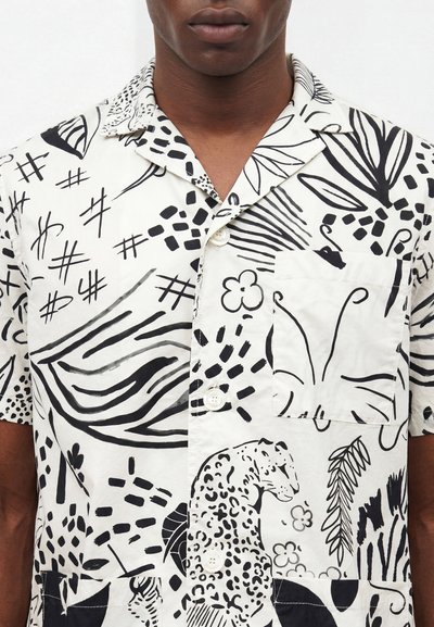 Άνδρας που φοράει πουκάμισο με κοντά μανίκια και μαύρα αφηρημένα, ζωικά και φυτικά μοτίβα σε λευκό φόντο.