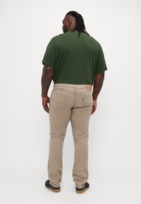 Pantalons tapered beige clair avec cinq poches, en tissu coton, et une patch en cuir à la taille arrière. Associé à une chemise à manches courtes vert foncé.