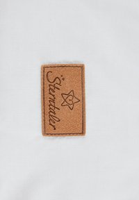 Braunes Lederlabel mit genähten Kanten, das den Text "Sterntaler" und ein Stern-Design zeigt, auf einem weißen Stoffhintergrund.