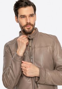 Giubbotto in pelle marrone con colletto alto, zip frontale e tasche con zip. Texture liscia e leggera lucentezza, cuciture dettagliate.