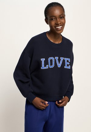 Smiling woman wearing a navy sweatshirt with "LOVE" in blue letters, paired with matching blue pants, standing against a plain background.