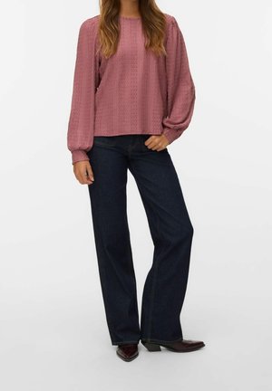Femme portant un chemisier texturé rose poudré à manches longues bouffantes, jean large foncé et chaussures foncées à bouts pointus, debout sur fond blanc.