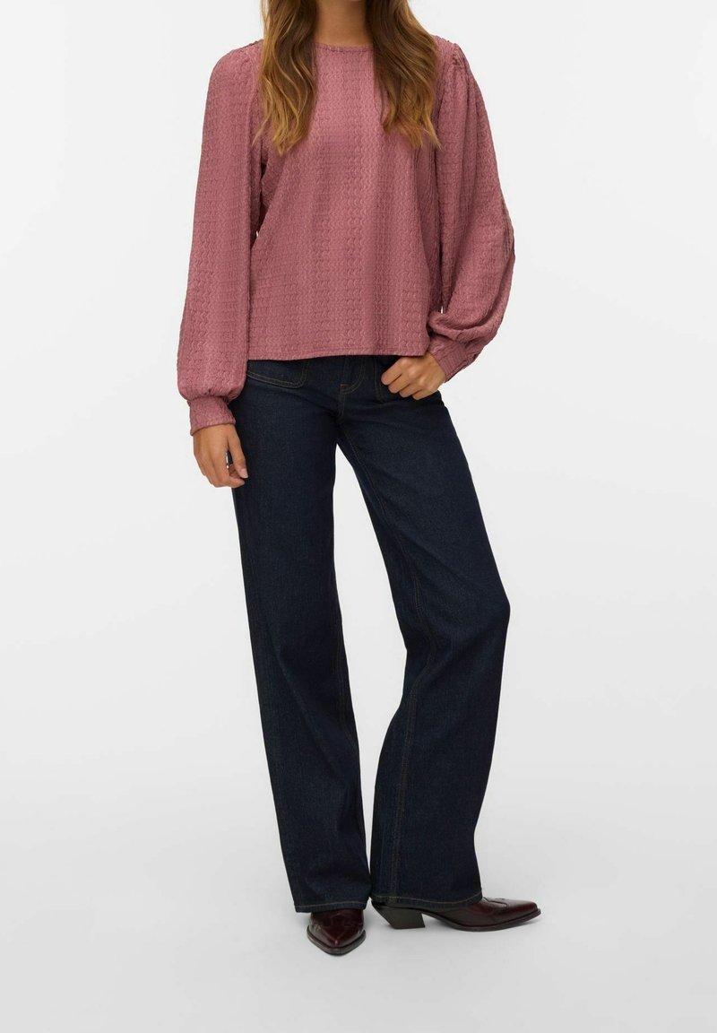 Femme portant un chemisier texturé rose poudré à manches longues bouffantes, jean large foncé et chaussures foncées à bouts pointus, debout sur fond blanc.