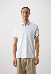 Camisa polo blanca con cuello a rayas en negro y azul marino, de manga corta y un logo sutil en el pecho; combinada con pantalones ligeros color beige.