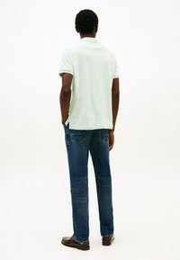 Lichtgroene polo met korte mouwen, gecombineerd met donkerblauwe jeans en bruine schoenen, gezien vanuit de achterkant tegen een effen achtergrond.