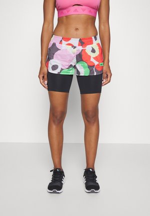 Femme portant un short fleuri coloré avec un short de compression noir intégré, un brassard de sport rose, et des baskets noires Adidas, debout sur fond blanc.