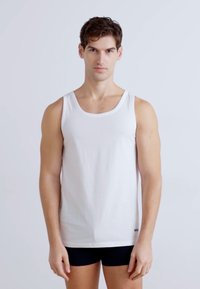 Baltas kokvilnas tank top ar apaļu kakla izgriezumu, pieguļoša forma un bez rakstiem. Modelis valkā melnus šortus, stāvot pret vieglu fonu.