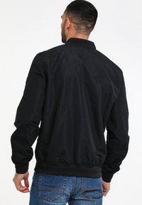 Sort bomberjakke med ribbet krave og manchetter, præget af en glat, mat tekstur og elastisk kant, båret over blå jeans.