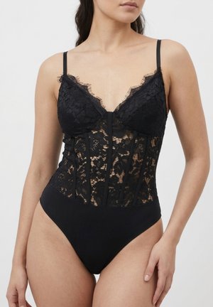 Mujer vistiendo un body de encaje negro con patrón floral translúcido y tirantes finos, posando frente a un fondo claro y liso.