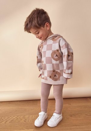 Jong kind in beige en witte geruite trui met teddybeer gezichten, beige legging en witte sneakers staand op een houten vloer.