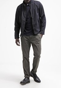 Giacca nera con colletto sopra una camicia scura, pantaloni khaki e scarpe nere stringate. Tessuto liscio con un design aderente e orli dei pantaloni arrotolati.