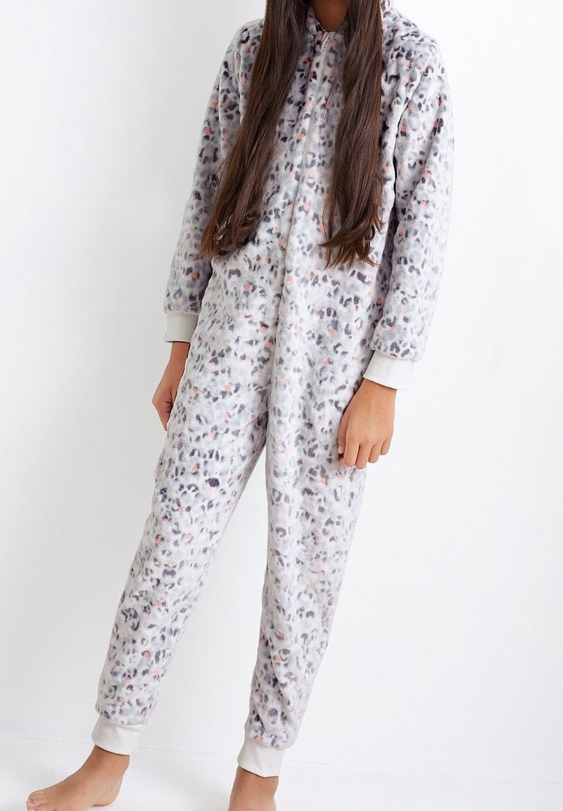 Personne portant une grenouillère en polaire à manches longues, douce, à imprimé léopard couvrant tout le corps, avec des poignets blancs, debout pieds nus sur un fond uni.