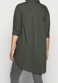 Olive green tunic shirt with a collared neck, half sleeves, side slits, and a curved hem. Made from lightweight fabric with a smooth texture.