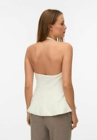 Bela peplum majica s halter vratnim dizajnom, prilagođena korzetu, gladke teksture, s blagim naborom na struku, u kombinaciji s bež hlačama.