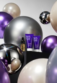 Trois tubes de soins NIP+FAB violets et une bouteille de sérum doré sont exposés sur un piédestal noir, entourés de ballons argentés, violets et beiges.