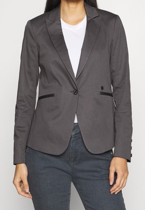 Femme portant un blazer gris ajusté sur une chemise blanche et un pantalon foncé, debout devant un fond clair uni.