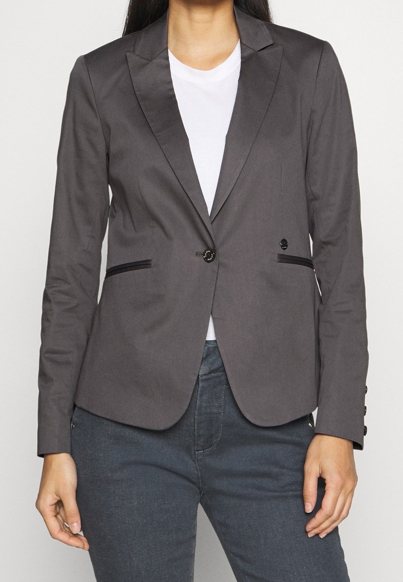 Femme portant un blazer gris ajusté sur une chemise blanche et un pantalon foncé, debout devant un fond clair uni.
