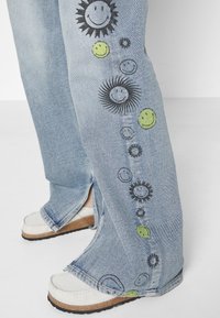 Jeans de mezclilla azul claro con estampados de cara sonriente y sol a lo largo del costado. Presenta un diseño de pierna ancha y dobladillo dividido, combinados con zapatos blancos.