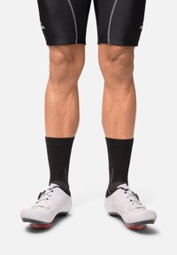 Zapatos de ciclismo blancos con suelas negras, calcetas negras y pantalones cortos negros. Los zapatos tienen una superficie texturizada, cordones y orificios de ventilación.