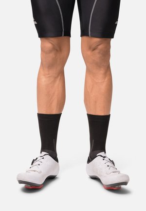 Chaussures de cyclisme blanches avec semelles noires, chaussettes noires et shorts noirs. Les chaussures présentent une surface texturée, des lacets et des trous de ventilation.