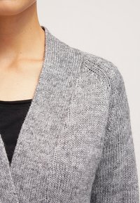 Záběr zblízka na osobu, která má na sobě šedou pletenou kardiganu přes černý top, ukazující texturu a detail na ramenou.