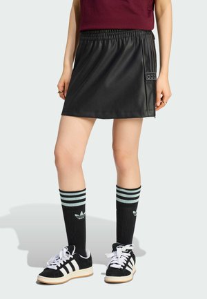 Persoon draagt een zwarte minirok van nepleer, zwarte Adidas kniekousen met lichtblauwe strepen en zwarte Adidas sneakers met witte strepen.