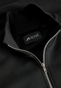 Black zip-up jacket with a high collar, ribbed cuffs, and a silver zipper. Features a textile label with the word "aprel." Soft fabric texture.