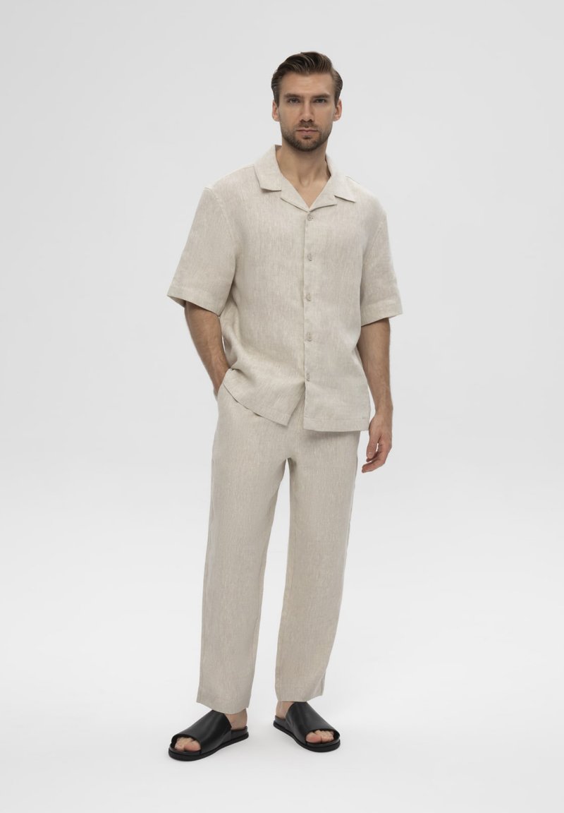 Homme portant une chemise beige à manches courtes boutonnée et un pantalon assorti avec des sandales noires ouvertes, debout avec une main dans la poche sur fond uni.
