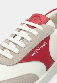 Άσπρο και μπεζ αθλητικό παπούτσι με κόκκινες λεπτομέρειες και λευκά κορδόνια, διακοσμημένο με κόκκινο λογότυπο και την επιγραφή "VALENTINO" στην πλευρική επιφάνεια.