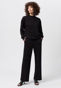 Top nero a maniche lunghe con collo alto e pantaloni neri a gamba larga. Tessuto morbido, vestibilità rilassata, abbinato a sandali neri con punta aperta.