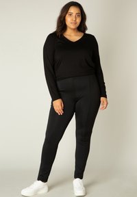 Zwarte langmouw V-hals top gecombineerd met zwarte leggings, beide gemaakt van een gladde stof. Model draagt witte sportschoenen.