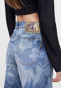 Γυναίκα που φοράει μπλε τζιν acid-wash με ετικέτα Just Cavalli και μια μαύρη κοντή μπλούζα με μακριά μανίκια που δείχνει μέρος της πλάτης της.