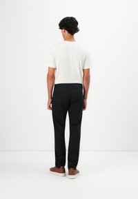Mand med mørkt krøllet hår iført hvid t-shirt, sorte bukser og brune sneakers står overfor en hvid væg med hænderne ned langs siden.