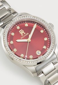 Stříbrné kovové hodinky s červeným ciferníkem, zlatými akcenty, markery připomínajícími diamanty a řemínkem posetým krystaly. Obsahují také zobrazení data.