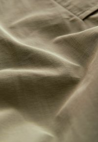 Tessuto leggero e strutturato in un tono khaki tenue, caratterizzato da morbide pieghe e una lucentezza sottile, con sottili linee orizzontali visibili in tutta la superficie.
