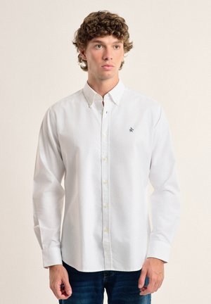 Camisa blanca de botones con mangas largas, hecha de una tela suave. Presenta un pequeño logo en azul marino y botones amarillos. Combinada con jeans de mezclilla azules.