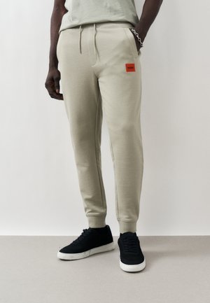 Man wearing light beige jogger pants with red Hugo patch, black sneakers with white soles, and a silver chain bracelet on left wrist.
