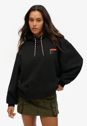Black hooded sweatshirt with a front pocket, featuring red logo text and patterned drawstring. Paired with an olive green cargo skirt.
