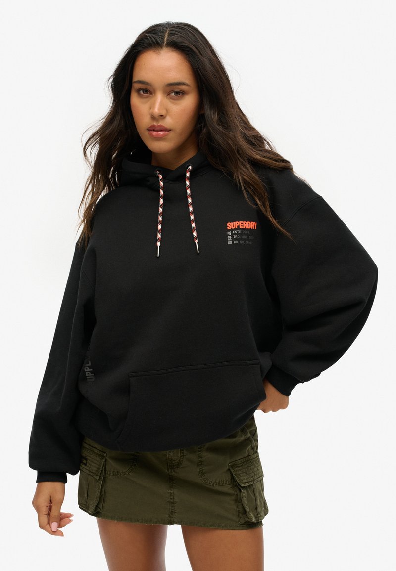 Sweat-shirt à capuche noir avec une poche avant, arborant un texte logo rouge et un cordon de serrage à motifs. Associé à une jupe cargo vert olive.