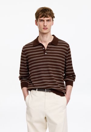 Jeune homme portant un polo à manches longues rayé marron et blanc et un pantalon beige, les mains dans les poches, sur un fond blanc uni.